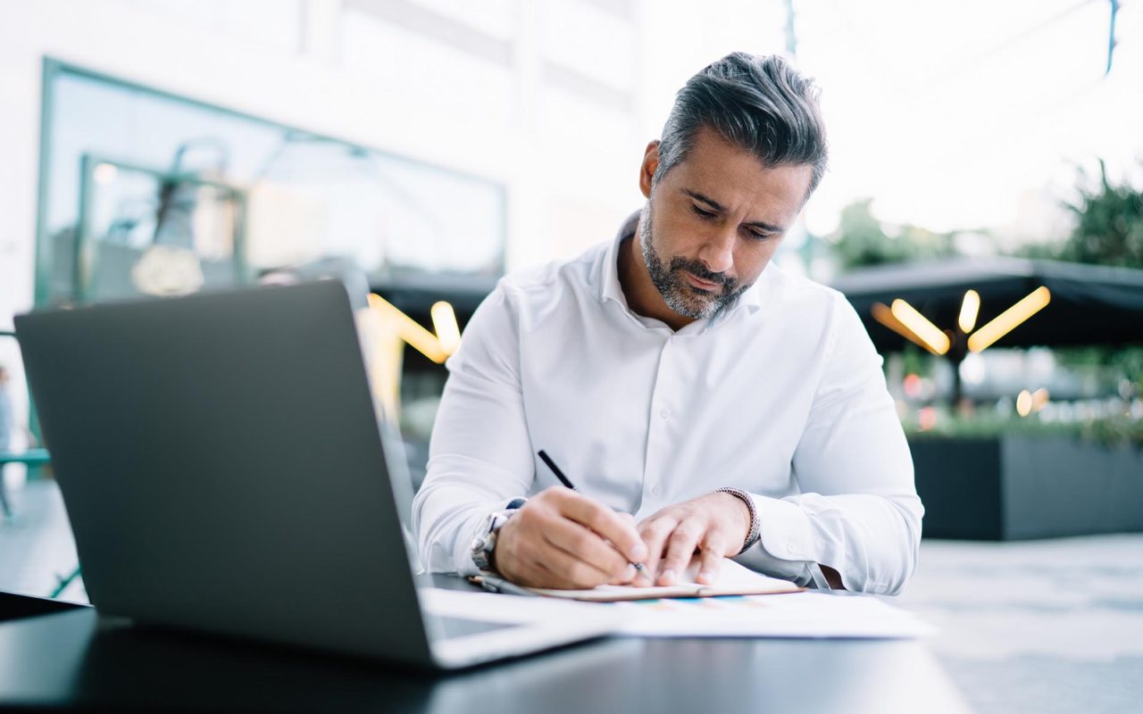 Mature entrepreneur writing informative notes in textbook sitting in sidewalk cafeteria with modern netbook and working remotely, middle aged proud CEO planning business meeting in financial district