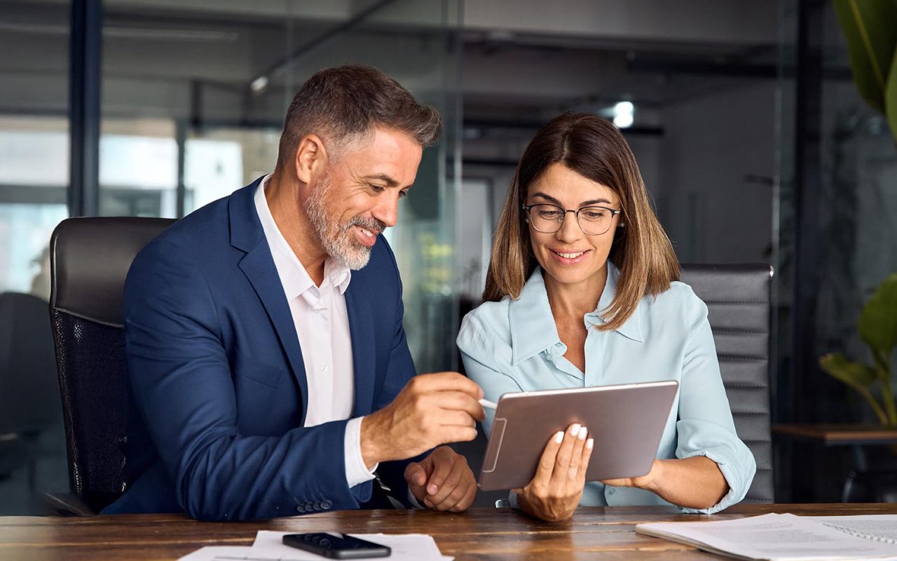 Two colleagues, group of partners, professional people working together. Mature 50s age Latin business man mentoring mid age European woman discussing project on tablet in office. Banner, copy space