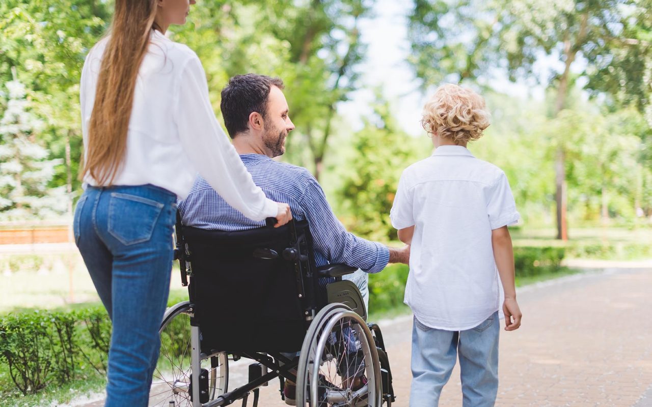 back view of mother rolling wheelchair with disabled father in park